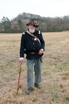 An elderly man wearing a historical military uniform stands in an open field. He is dressed in a dark blue jacket with gold buttons and epaulettes, light blue pants, and a hat adorned with a badge. He holds a wooden walking stick and has a beard. The background features a landscape with grass and a line of trees under a cloudy sky.