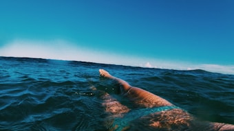 A person is swimming in open water, with their arm reaching forward. The surface of the water is slightly rippled, reflecting sunlight, under a clear blue sky.