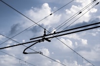 Overhead electrical wires crisscross against a backdrop of a cloudy sky, with various cables and connectors visible.