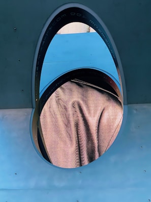 An oval-shaped digital display is embedded in a wall. The screen shows a close-up view of a fabric texture in a peachy color. Surrounding the display is a metallic border with inscriptions. The wall is a smooth, flat surface in a muted blue tone, with a few visible screws or bolts.