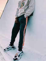A person wearing a light-colored hoodie and dark sweatpants with orange stripes, leaning against a wall. They are also wearing black and white sneakers with prominent designs and have a gold watch. The setting appears to be outdoors with a white, textured background.