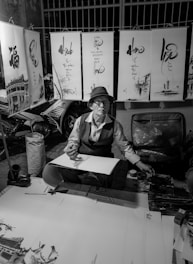 An artist is seated at a small table surrounded by calligraphy artworks hanging above. Wearing a hat and glasses, the artist appears focused while holding a brush poised over a partially completed piece. The environment suggests a street market or outdoor setting, likely illuminated by artificial light. Various tools and materials are scattered around, indicating an active workspace for creating art.