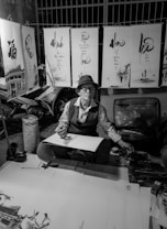 An artist is seated at a small table surrounded by calligraphy artworks hanging above. Wearing a hat and glasses, the artist appears focused while holding a brush poised over a partially completed piece. The environment suggests a street market or outdoor setting, likely illuminated by artificial light. Various tools and materials are scattered around, indicating an active workspace for creating art.