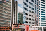 A cityscape featuring several tall modern skyscrapers with a few recognizable logos on their facades. The buildings are closely situated, showcasing a mix of glass and concrete architectural designs. A construction crane is visible among the buildings, indicating ongoing development in the area.