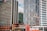 A cityscape featuring several tall modern skyscrapers with a few recognizable logos on their facades. The buildings are closely situated, showcasing a mix of glass and concrete architectural designs. A construction crane is visible among the buildings, indicating ongoing development in the area.