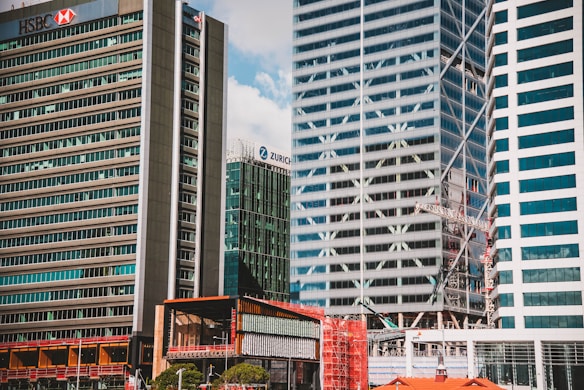 A cityscape featuring several tall modern skyscrapers with a few recognizable logos on their facades. The buildings are closely situated, showcasing a mix of glass and concrete architectural designs. A construction crane is visible among the buildings, indicating ongoing development in the area.