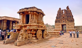 A remarkable stone chariot is prominently displayed in an ancient temple complex, accompanied by intricately carved details and sculpture designs. Several people are gathered around, either observing or taking photographs. In the background, a towering, weathered entrance structure is visible, contributing to the historical ambiance of the scene.
