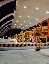 A modern toll plaza is seen at night, featuring a well-lit arched canopy structure with multiple toll booths lined up. Several vehicles are positioned at the booths, including a white and a black car. The area is illuminated by overhead lights, creating a bright and organized atmosphere. Potted plants hang from yellow and red barriers delineating lanes.