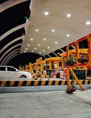 A modern toll plaza is seen at night, featuring a well-lit arched canopy structure with multiple toll booths lined up. Several vehicles are positioned at the booths, including a white and a black car. The area is illuminated by overhead lights, creating a bright and organized atmosphere. Potted plants hang from yellow and red barriers delineating lanes.