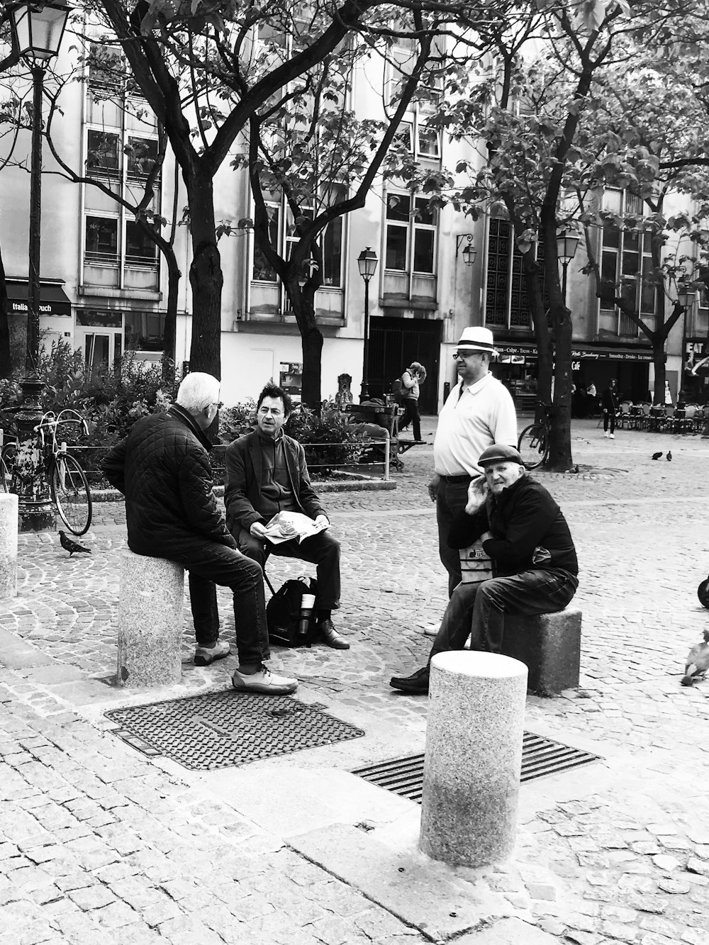 three men sitting and one man standing on concrete road beside trees and buildings