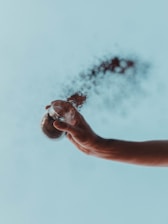A hand is holding a small glass jar from which black powder or dust is being released and disperses into the air against a light blue sky background.