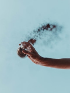 A hand is holding a small glass jar from which black powder or dust is being released and disperses into the air against a light blue sky background.