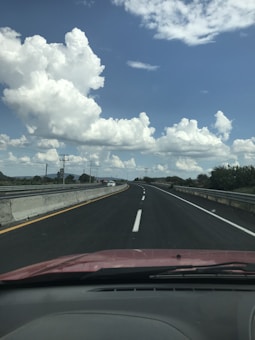 A highway stretches into the distance under a sky filled with large, fluffy clouds. The road is clean and well-maintained, lined with concrete barriers on either side. A few vehicles can be seen on the road, and the scenery includes trees and telephone poles on the horizon.