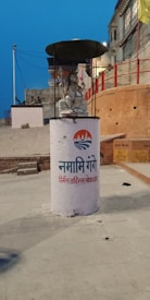 A statue of a seated figure under a black umbrella-like canopy is placed on a cylindrical pedestal with 'Namami Gange' written on it in Hindi. The background features a slightly inclined street with red-painted walls and buildings. The scene is set during the evening, as indicated by the blue sky.