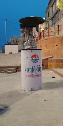 A statue of a seated figure under a black umbrella-like canopy is placed on a cylindrical pedestal with 'Namami Gange' written on it in Hindi. The background features a slightly inclined street with red-painted walls and buildings. The scene is set during the evening, as indicated by the blue sky.