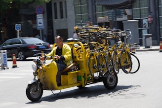 A man rides a yellow motorbike on a street, transporting several yellow bicycles on a specialized rack. The setting appears to be urban with cars and buildings in the background. Traffic signs and road markers are visible, suggesting an active city environment.