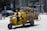 A man rides a yellow motorbike on a street, transporting several yellow bicycles on a specialized rack. The setting appears to be urban with cars and buildings in the background. Traffic signs and road markers are visible, suggesting an active city environment.