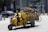 A man rides a yellow motorbike on a street, transporting several yellow bicycles on a specialized rack. The setting appears to be urban with cars and buildings in the background. Traffic signs and road markers are visible, suggesting an active city environment.