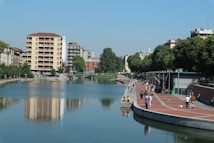 A scenic urban waterfront with a calm body of water reflecting nearby buildings. A pathway lined with trees and lampposts runs alongside, where people stroll and sit. The skyline is dotted with mid-rise buildings, blending architectural styles surrounded by greenery.