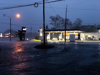A dimly lit gas station is seen during heavy rainfall. The pavement is wet and reflective, with puddles forming. The station's lights illuminate the area, creating a stark contrast with the dark, cloudy sky. There are cars parked at the pumps and signs displaying the station's brand.