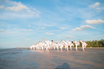 A group of people practicing martial arts on a sandy beach under a clear blue sky. They are lined up in formation, performing synchronized high kicks. The scene is peaceful with a vast expanse of open sky and a few scattered clouds. In the background, there are trees lining the edge of the beach.