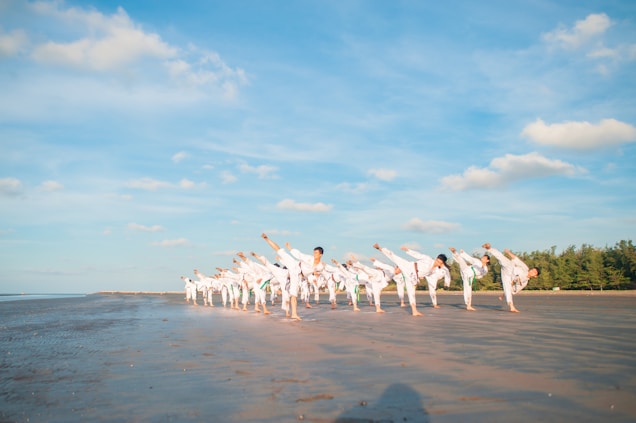 A group of people practicing martial arts on a sandy beach under a clear blue sky. They are lined up in formation, performing synchronized high kicks. The scene is peaceful with a vast expanse of open sky and a few scattered clouds. In the background, there are trees lining the edge of the beach.