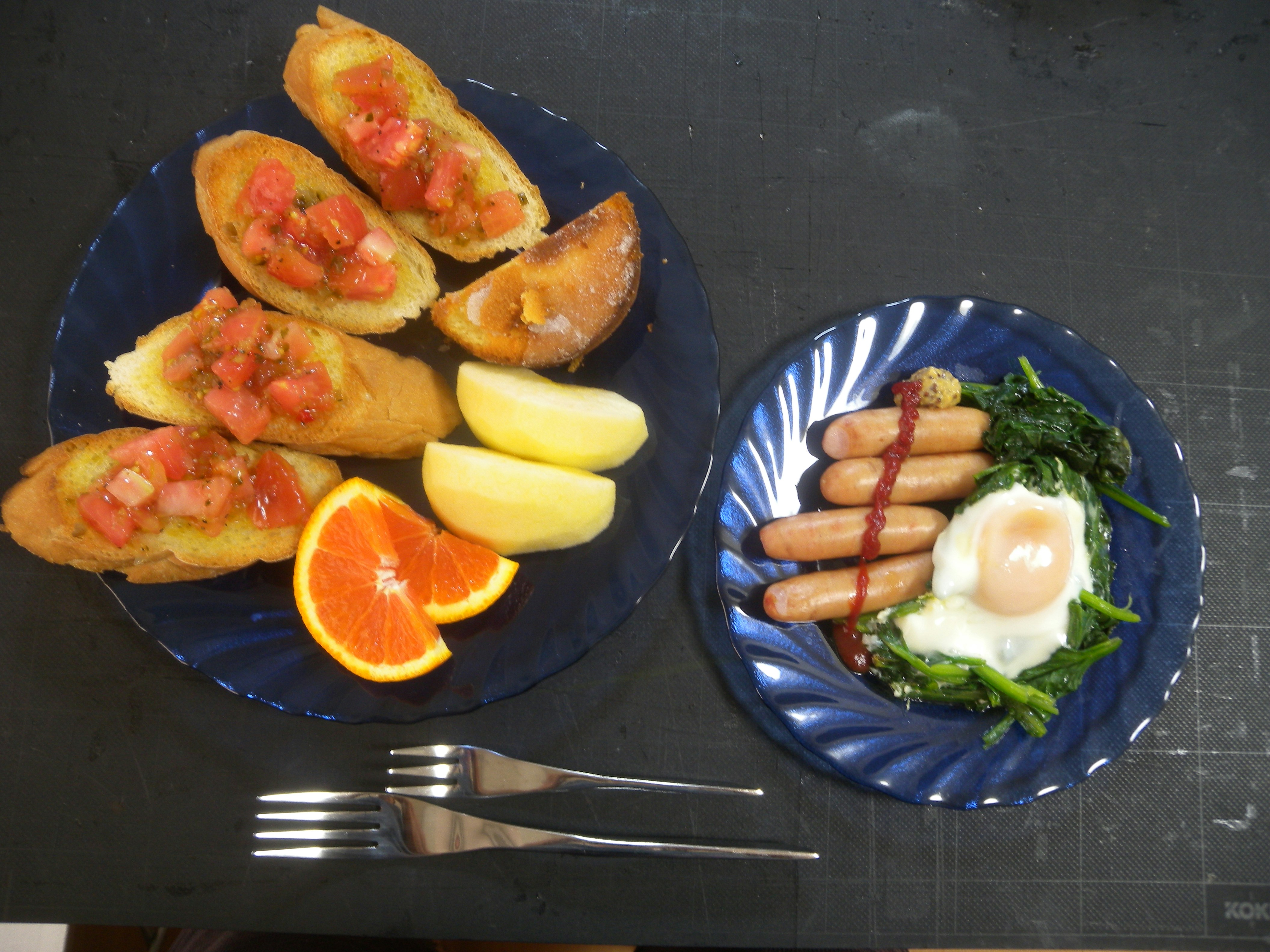 A meal is arranged on two blue plates. The left plate features four slices of bread topped with diced tomatoes, alongside quartered apple slices and an orange wedge. The right plate contains four sausages drizzled with sauce, accompanied by a sunny-side-up egg and sautéed greens. Two forks are positioned between the plates.