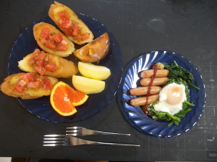 A meal is arranged on two blue plates. The left plate features four slices of bread topped with diced tomatoes, alongside quartered apple slices and an orange wedge. The right plate contains four sausages drizzled with sauce, accompanied by a sunny-side-up egg and saut&eacute;ed greens. Two forks are positioned between the plates.