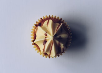 A single cupcake with smooth, cream-colored frosting topped with chocolate drizzle, viewed from above on a light background. The frosting is swirled in an intricate pattern, and the cupcake is encased in a yellow paper liner.