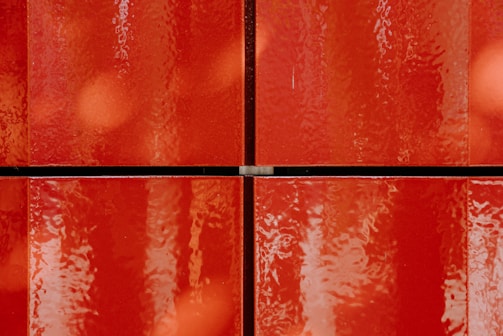 Four glossy red tiles with a slightly textured surface, arranged in a grid pattern. The tiles have a shiny, reflective quality, creating an impression of depth and richness.
