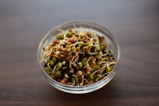 A clear glass bowl containing a mix of colorful sprouted legumes, including green, brown, and black seeds with visible white roots, is placed on a wooden surface.