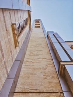 A tall building with a modern architectural design, featuring a combination of stone and metal panels. The perspective is angled upwards, emphasizing the height and structure of the building. Natural stone textures are visible along with reflective metal elements, and a series of rectangular windows is integrated into the design.