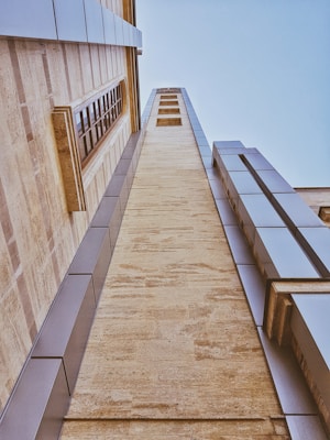 A tall building with a modern architectural design, featuring a combination of stone and metal panels. The perspective is angled upwards, emphasizing the height and structure of the building. Natural stone textures are visible along with reflective metal elements, and a series of rectangular windows is integrated into the design.