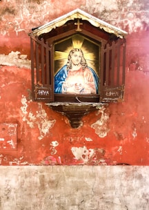 A religious icon encased in a decorative frame is mounted on an aged, weathered wall. The icon features a portrait of a figure with long hair and a halo, depicted holding a heart. The frame has a small roof with a cross on top, providing a modest shelter for the icon.