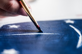 A close-up of a hand using a paintbrush to apply blue paint onto a surface, featuring white stars and stripes, suggesting a flag or patriotic theme.