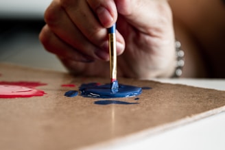 A close-up view of a hand holding a paintbrush, applying blue paint onto a surface. The hand appears focused, and there are visible splatters of blue and red paint on the surface.