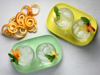 Two trays, one yellow and one green, each hold two glasses filled with ice and garnished with mint leaves, lemon, and orange slices. Adjacent to the trays, there is a collection of decorative orange peel spirals arranged artistically on a light surface.