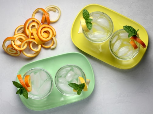 Two trays, one yellow and one green, each hold two glasses filled with ice and garnished with mint leaves, lemon, and orange slices. Adjacent to the trays, there is a collection of decorative orange peel spirals arranged artistically on a light surface.