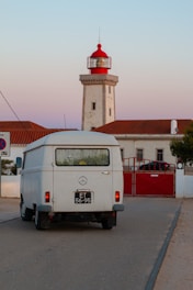 A vintage white van with a license plate is parked on a road near a red and white lighthouse. The lighthouse and adjacent building have red roofs and are set against a sky transitioning from pinkish hues to blue, suggesting sunset.