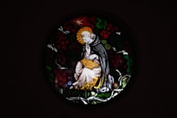 A stained glass window depicts a serene figure in religious robes, sitting thoughtfully with a halo around their head. The figure holds an open book, surrounded by a circular design of leaves and berries.