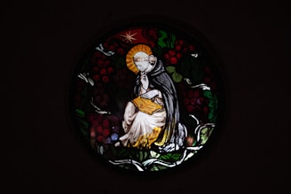 A stained glass window depicts a serene figure in religious robes, sitting thoughtfully with a halo around their head. The figure holds an open book, surrounded by a circular design of leaves and berries.