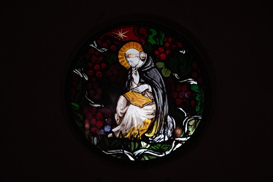 A stained glass window depicts a serene figure in religious robes, sitting thoughtfully with a halo around their head. The figure holds an open book, surrounded by a circular design of leaves and berries.