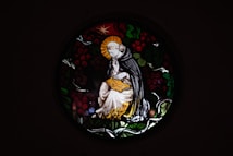 A stained glass window depicts a serene figure in religious robes, sitting thoughtfully with a halo around their head. The figure holds an open book, surrounded by a circular design of leaves and berries.