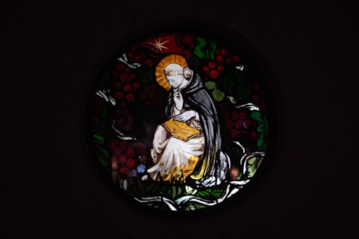 A stained glass window depicts a serene figure in religious robes, sitting thoughtfully with a halo around their head. The figure holds an open book, surrounded by a circular design of leaves and berries.