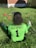 A person with curly hair sits on green grass facing away, wearing a bright green sports jersey with 'B & G SPORTS' and the number 1 on the back. Sports equipment, including a red helmet and baseballs, is scattered around.