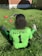 A person with curly hair sits on green grass facing away, wearing a bright green sports jersey with 'B & G SPORTS' and the number 1 on the back. Sports equipment, including a red helmet and baseballs, is scattered around.