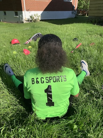 A person with curly hair sits on green grass facing away, wearing a bright green sports jersey with 'B & G SPORTS' and the number 1 on the back. Sports equipment, including a red helmet and baseballs, is scattered around.