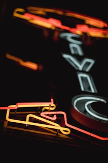 A neon sign is illuminated with vibrant colors against a dark background, featuring bold lines and shapes that give it a retro aesthetic. The glowing tubes form text and potential symbols, creating a striking visual effect.