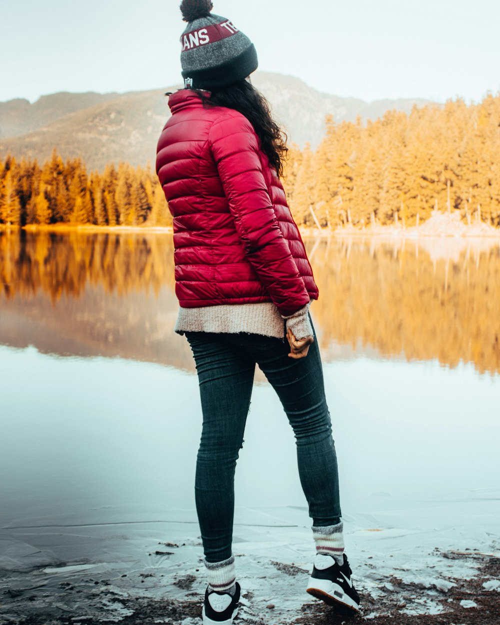 woman in red bubble jacket and blue skinny denim jeans standing in bank of lake