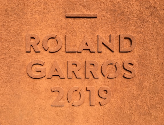Large engraved letters displaying the words 'Roland Garros 2019' are set into a textured clay surface, suggesting an association with the famous French Open tennis tournament.